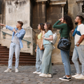 A guide points toward a historic building as a small group of people in casual attire look up, listening attentively.