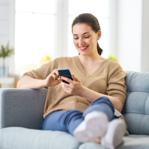 A person wearing a tan sweater smiles while relaxing on a blue couch and looking at a smartphone held in both hands.