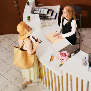 A visitor in a hat and yellow dress talks to a receptionist at a modern white hotel front desk.