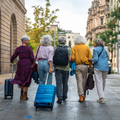 Five individuals walk together down a paved city street, carrying luggage and backpacks, viewed from behind.