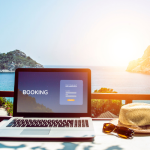 A laptop showing a booking screen sits on a table overlooking the ocean with a straw hat and sunglasses nearby.