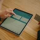 A person sitting at a wooden desk uses a tablet to make an online payment while holding a credit card.