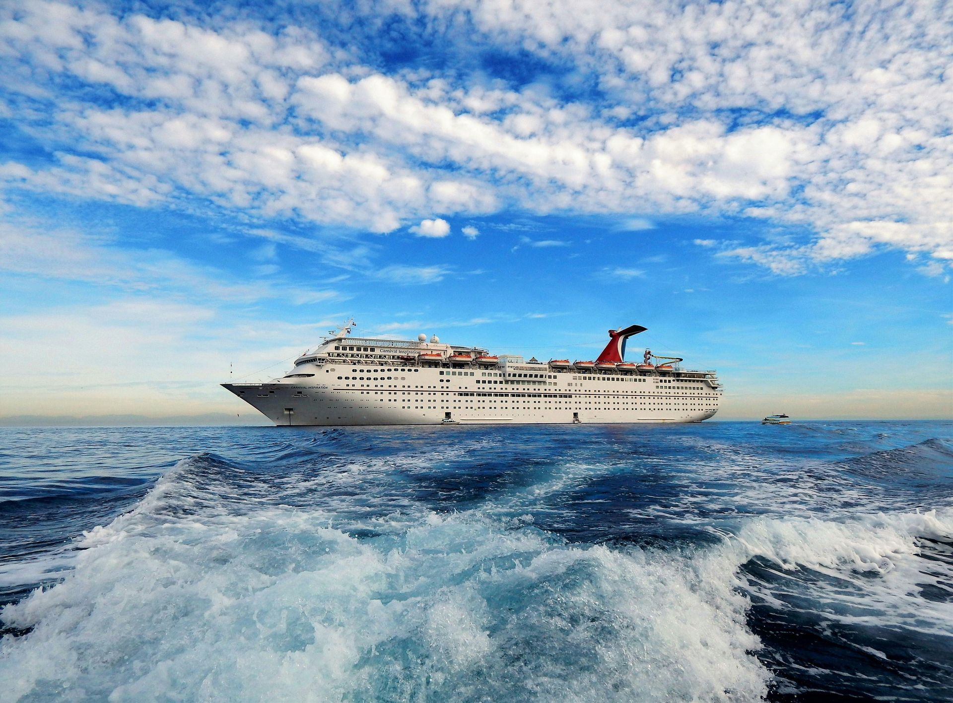 A large Carnival cruise ship on the open ocean under a bright blue, cloudy sky, seen from the ship's wake.