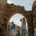 Three people walk through a stone archway in ancient, sunlit ruins set against a mountain landscape.