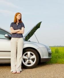 Woman standing with arms crossed next to a car with an open hood on a roadside.