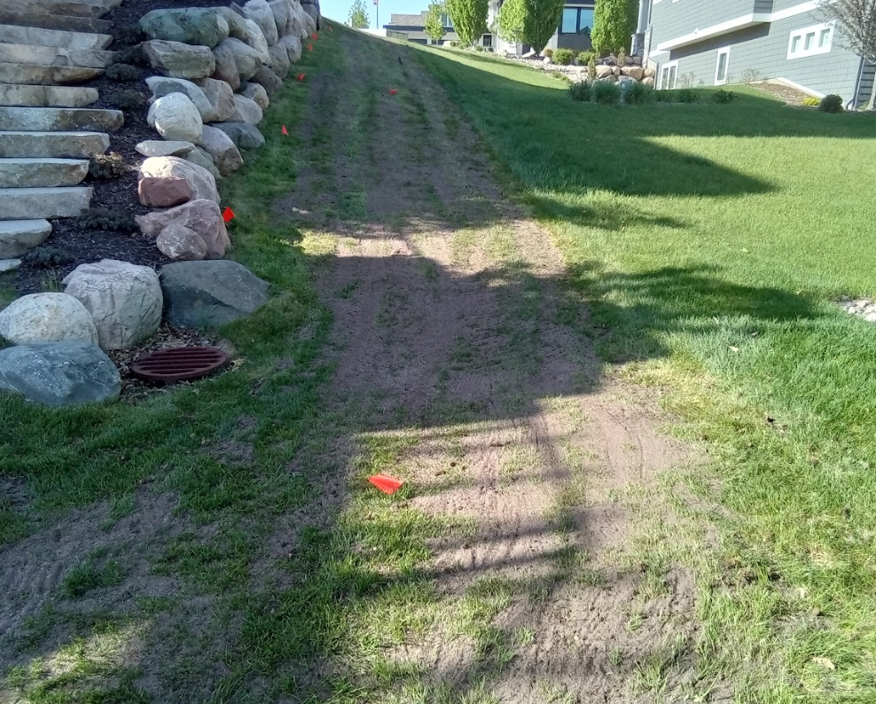 A sloped yard with a bare dirt path beside a stone retaining wall and a manicured green lawn.