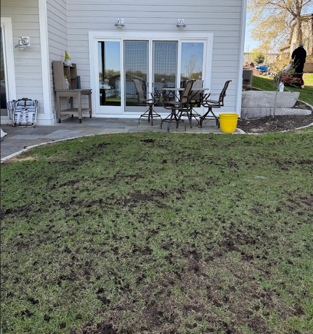 A gray house with a patio containing chairs and a table overlooking a lawn with patchy grass and exposed dirt.