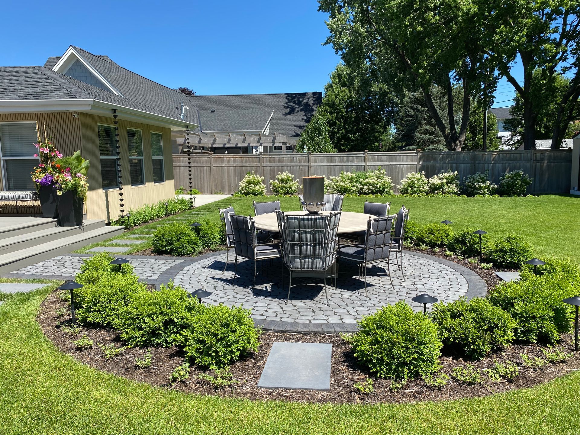 Cobblestone pavers backyard patio landscaping design KG landscape Edina MN inspiration ideas row of trees privacy cedar fence plantings table chairs stepping stones modern colorful boxwoods