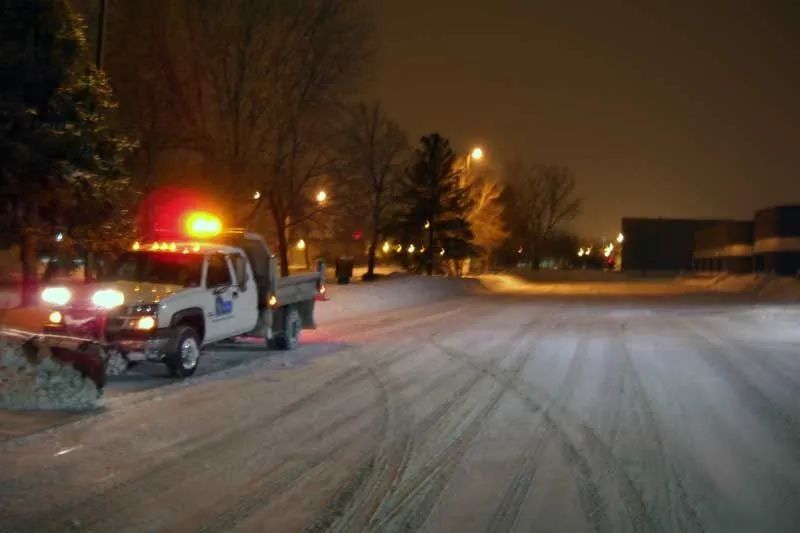 Snowplow clearing a snow-covered road lined with trees and bushes on a winter day.