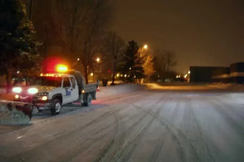 Snowplow clearing a snow-covered street at night, with flashing lights and streetlights illuminating the scene.