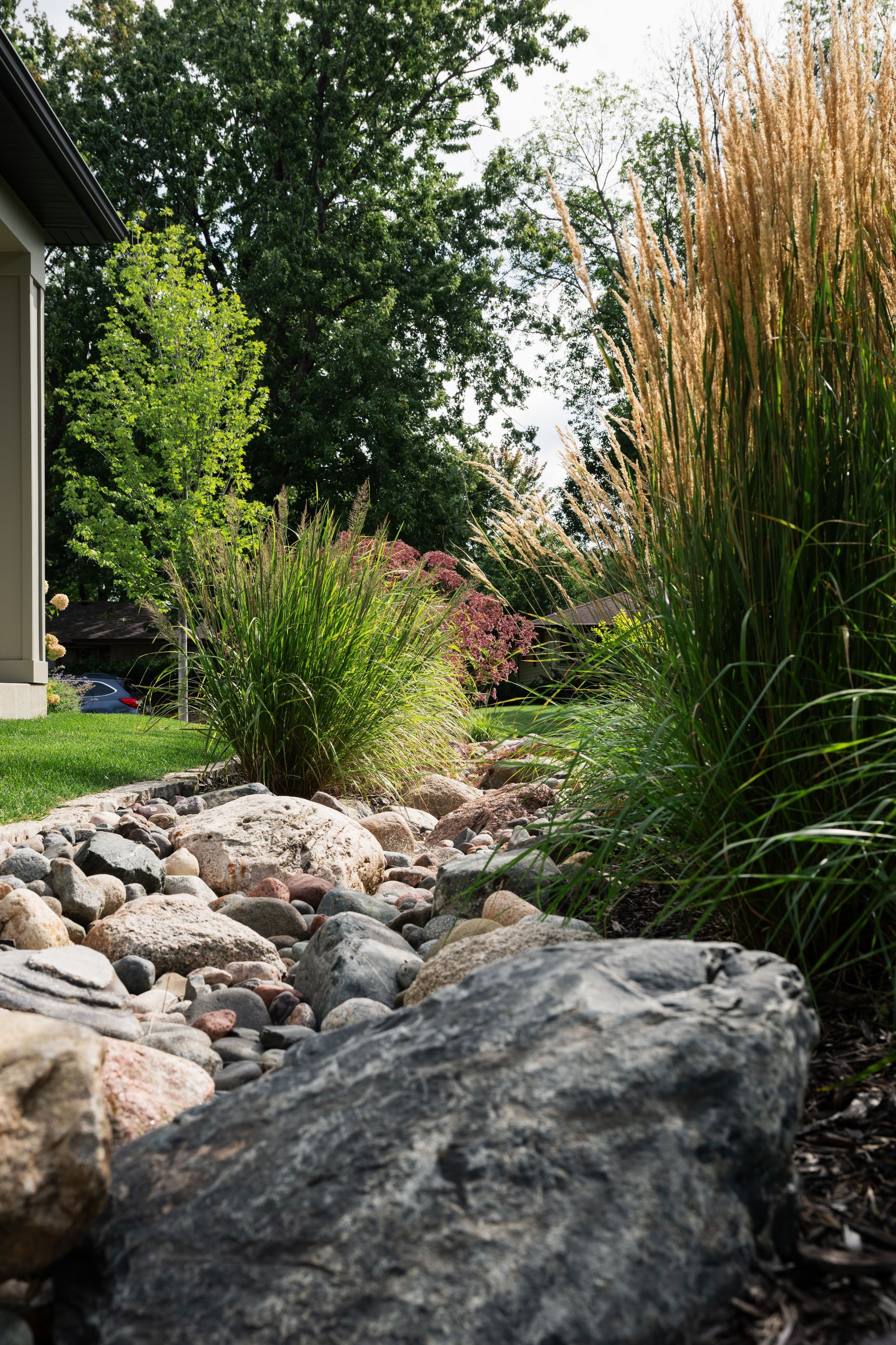 A house with a dry creek bed and colorful planting and nice landscaping is surrounded by trees and rocks in a garden.
