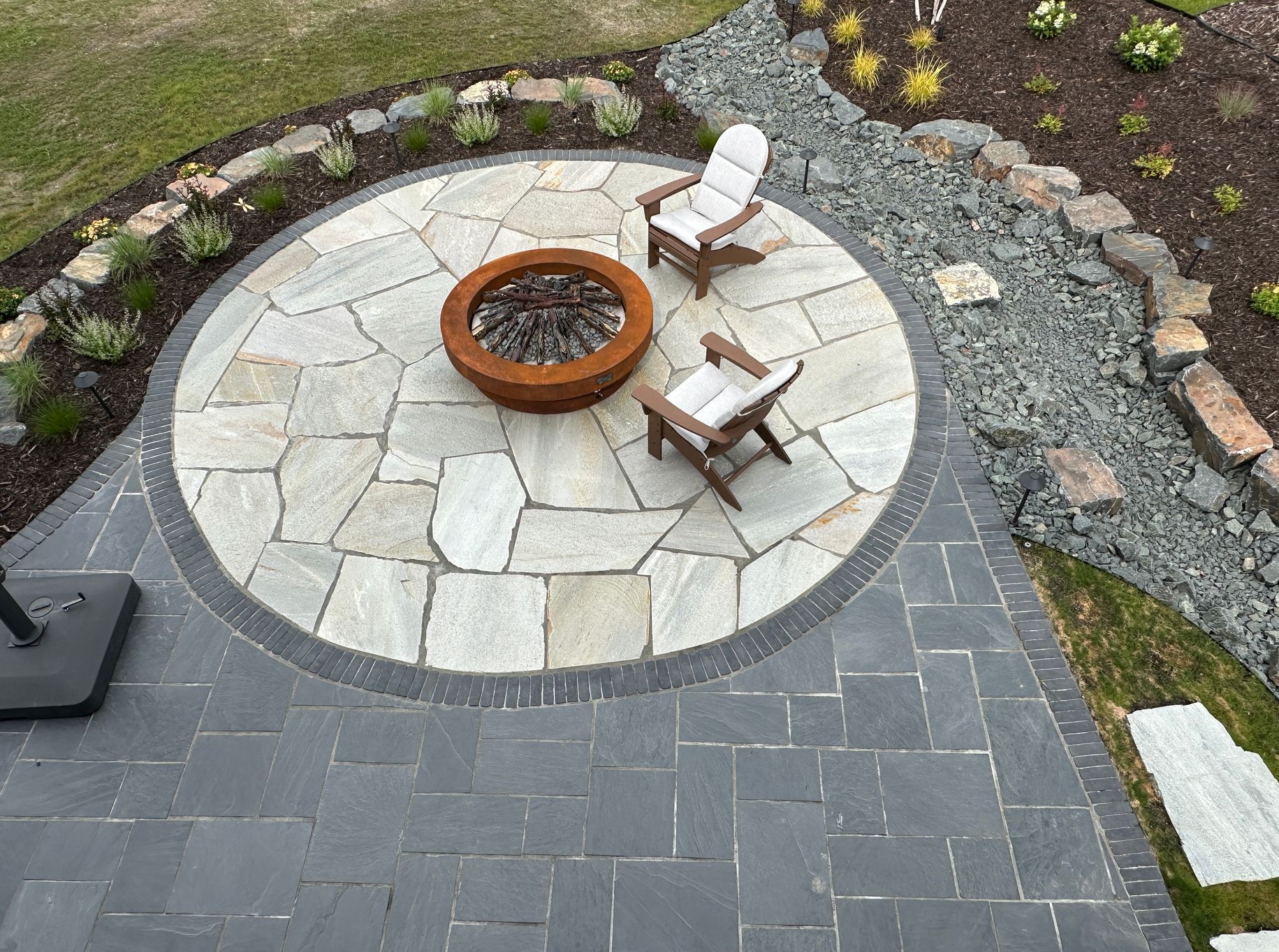 Raised patio round shape with gas fire feature built with large irregular gray silver flagstone with black paver border and dry creek bed KG Landscape MN 