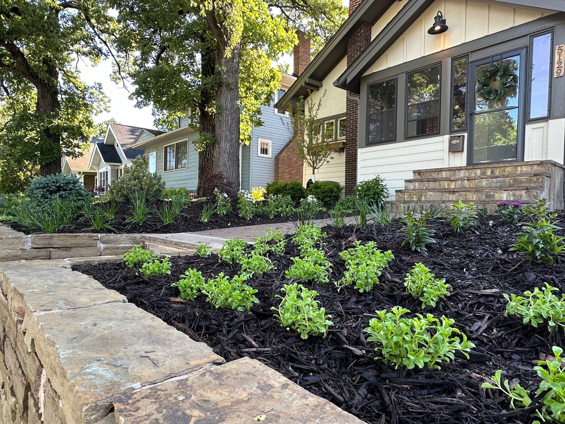 Stone-walled garden bed with young green plants in front of a white house with dark trim and a porch.
