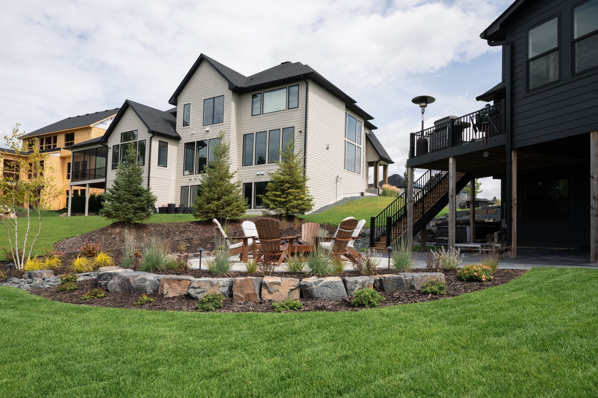 Raised circular flagstone patio with gas fire feature and Adirondack chairs with landscaping filled with boulders and flowers with thick green lawn in Plymouth MN 
