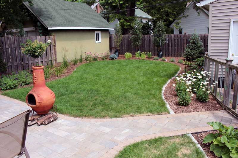 A backyard with a brick walkway leading to a house and a fence.