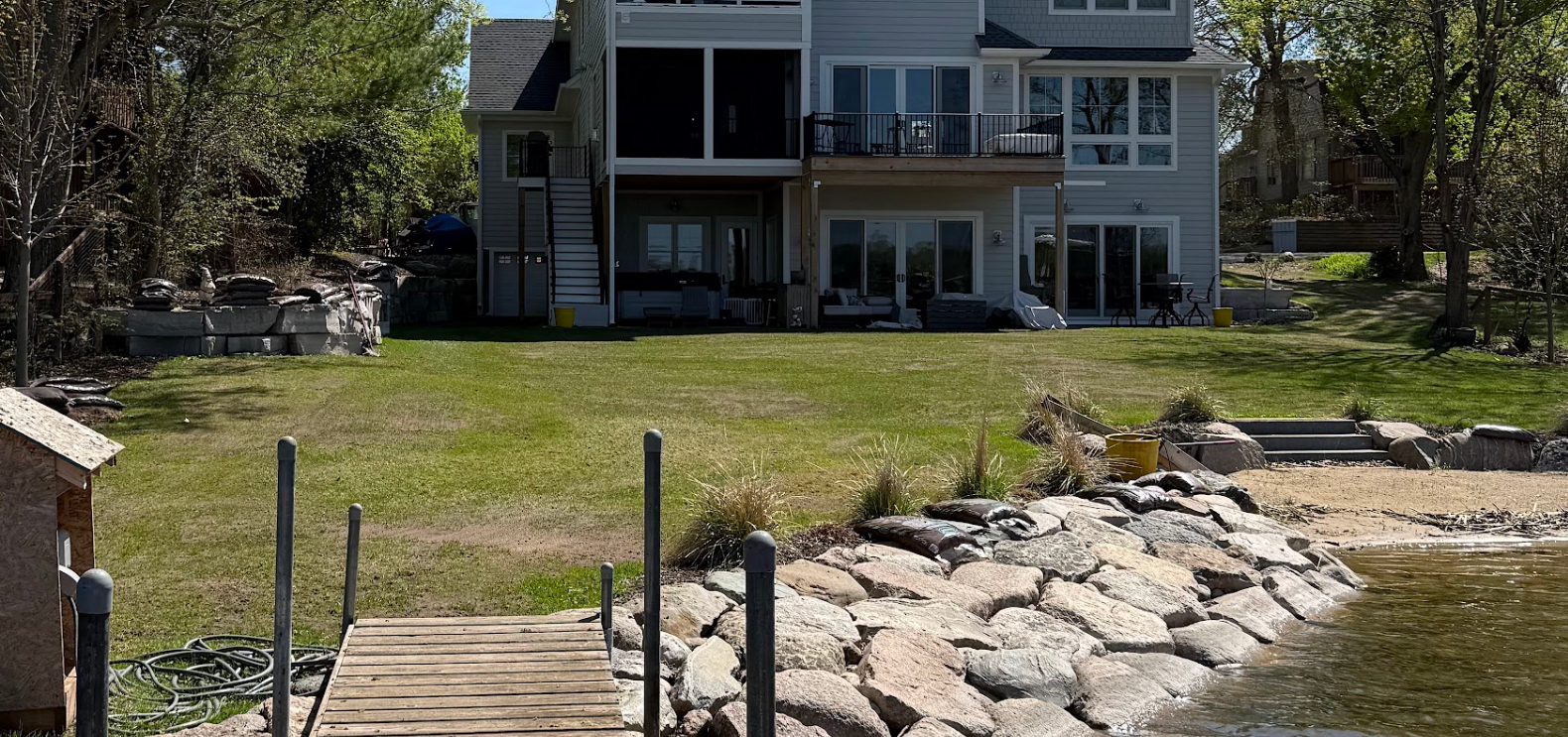 A two-story gray house sits on a grassy lawn behind a rocky shoreline and a wooden boat dock on a lake.