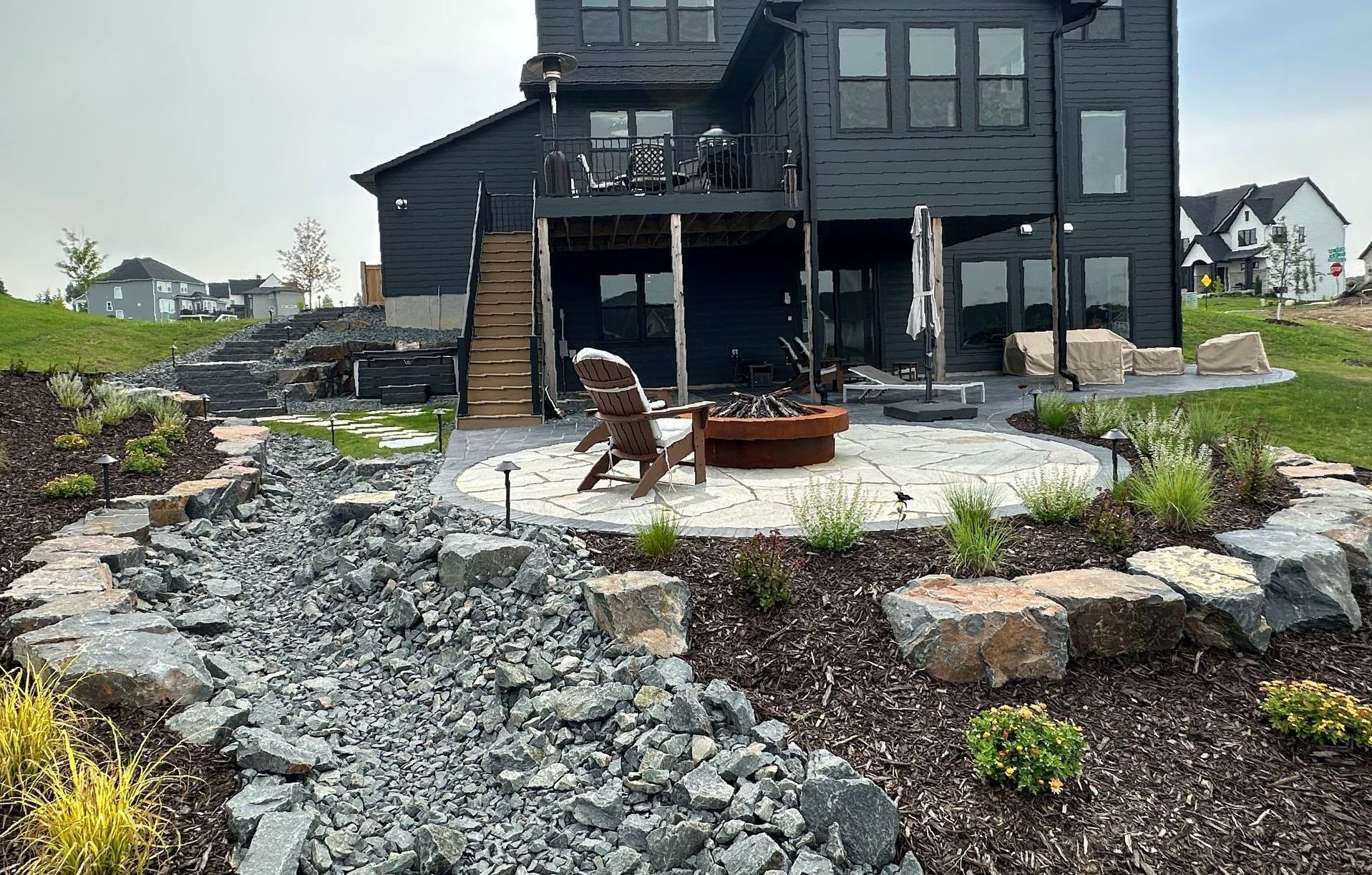 Dream backyard hardscaping and landscape design with flagstone patio off deck steps gas fire ring dry creek bed using Gray trap rock and boulders KG Landscape Plymouth MN