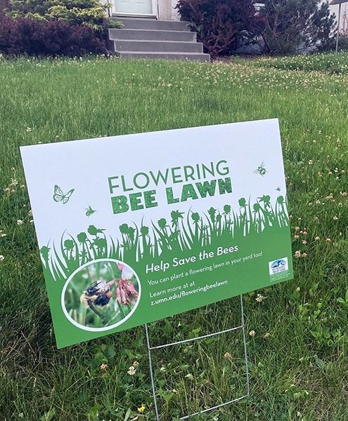 A flowering bee lawn sign is sitting in the grass in front of a house.