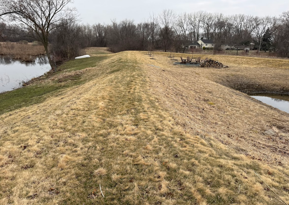 A grassy embankment slopes down to water on both sides under a cloudy sky, with trees and a distant house in the back.
