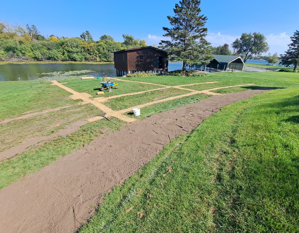 A landscape showing a lawn with freshly dug trenches and a curved dirt path leading toward two dark buildings by a lake.