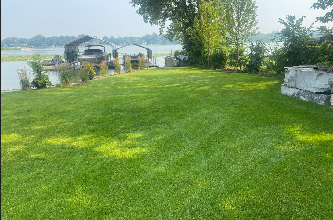 A lush green lawn leading to a waterfront with boats docked at a pier, framed by trees and a stone retaining wall.