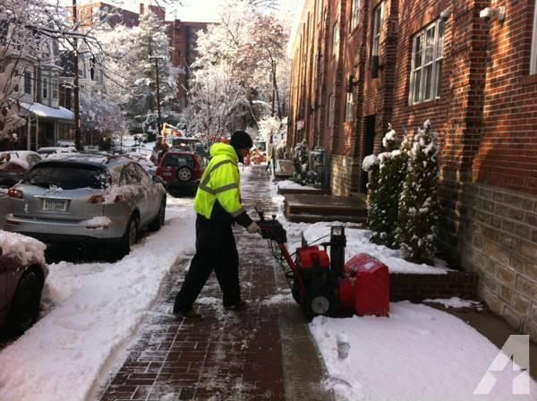 Person using a red snowblower on a snow-covered sidewalk, bordered by parked cars and a brick building.