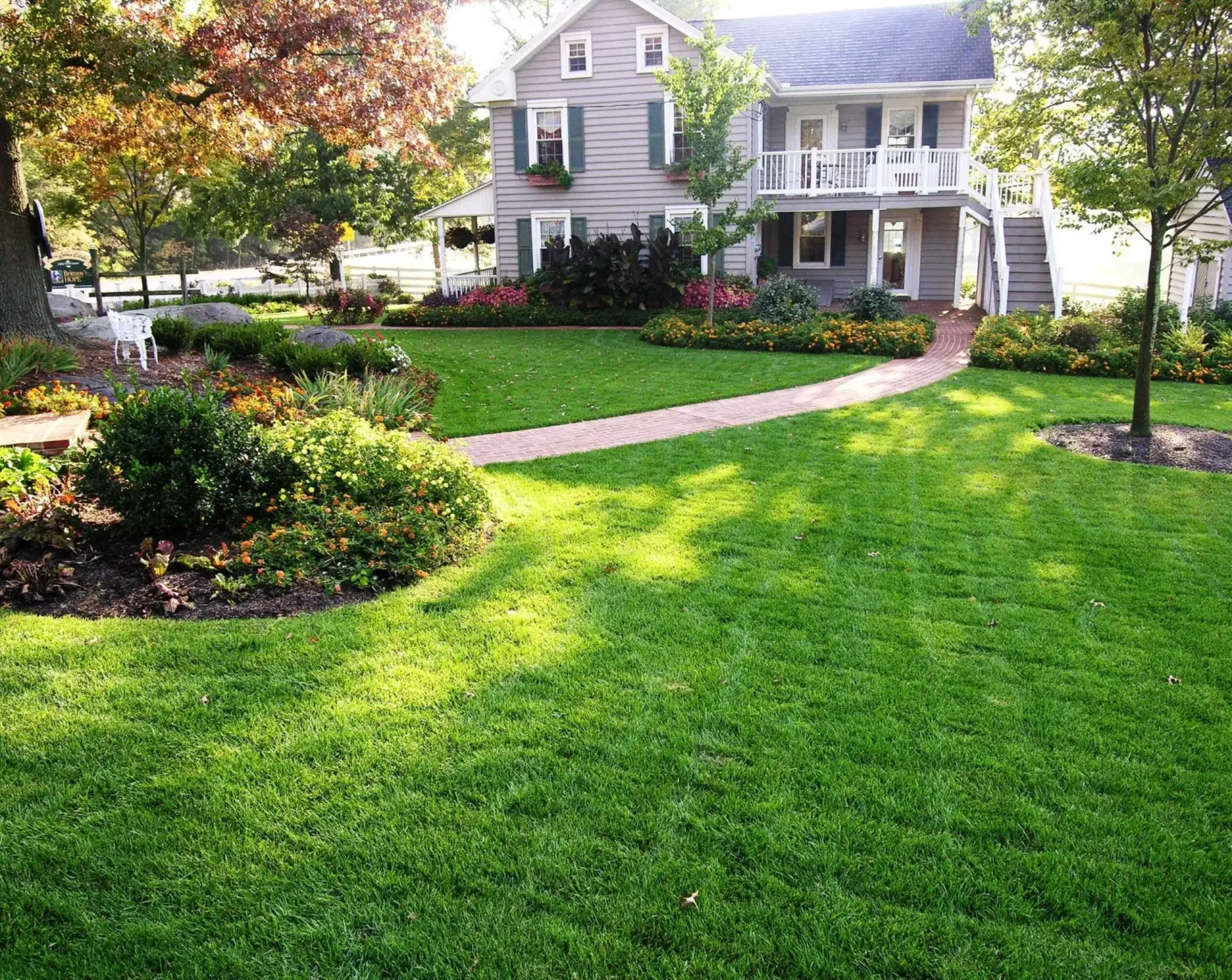 Lush green lawn with brick path leading to a gray house with a white balcony and colorful flower beds.