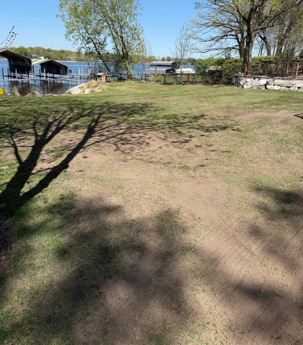 A view of a grassy yard leading toward a lake, featuring boat docks, shoreline rocks, and trees under a clear blue sky.