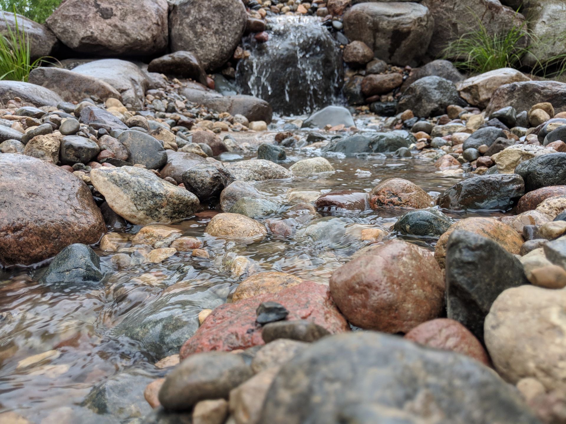 Stream flowing over rocks, small waterfall in background. Water is clear, rocks are various colors.