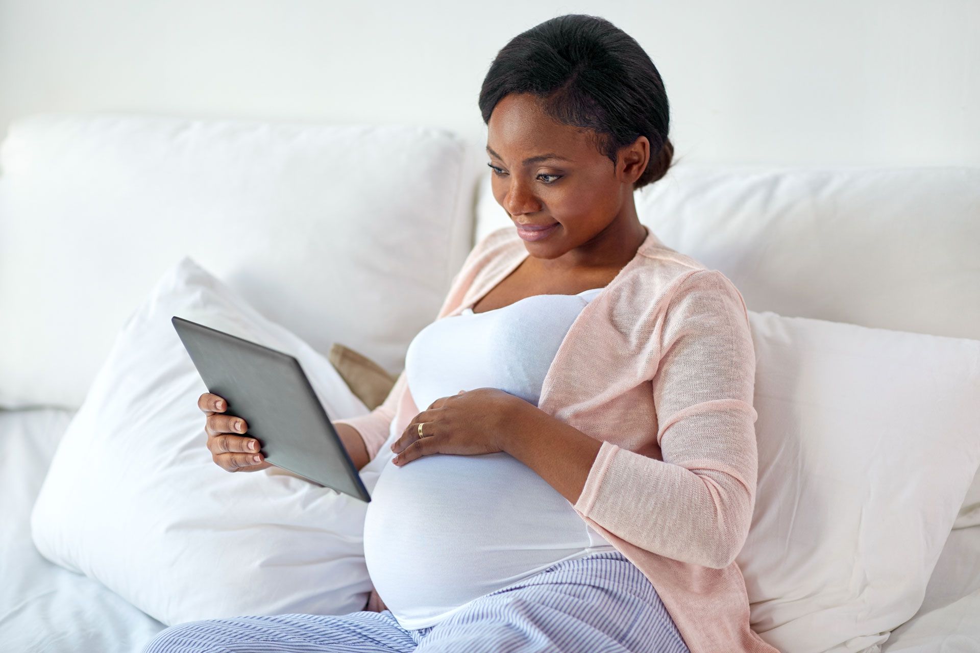 A pregnant woman is sitting on a bed using a tablet computer.