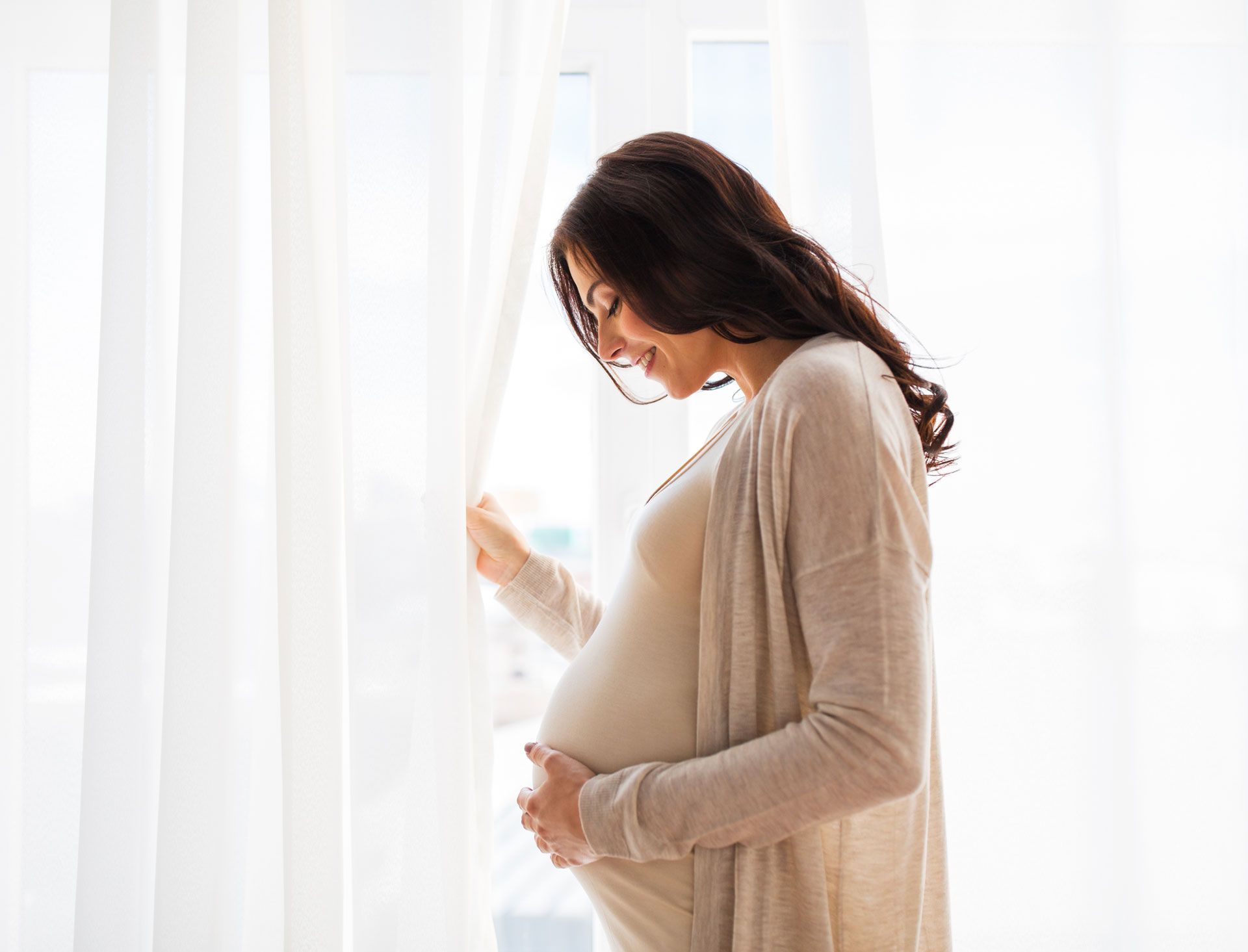 A pregnant woman is standing in front of a window holding her belly.