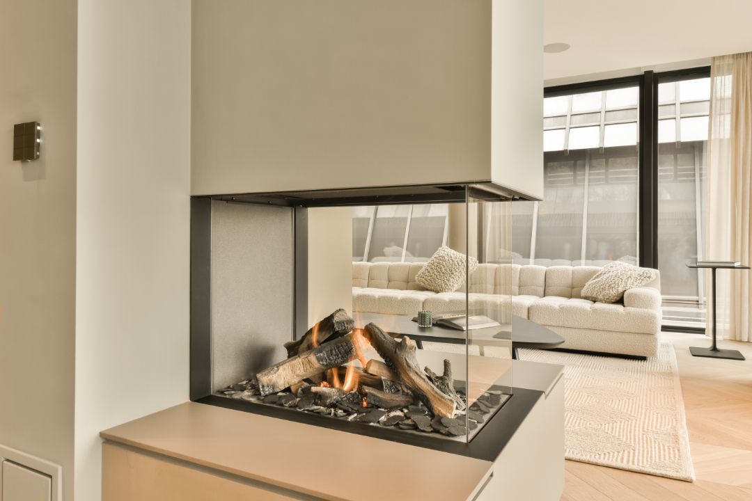 Modern fireplace with glass enclosure, burning faux logs. Beige walls, sofa, and light wood flooring.