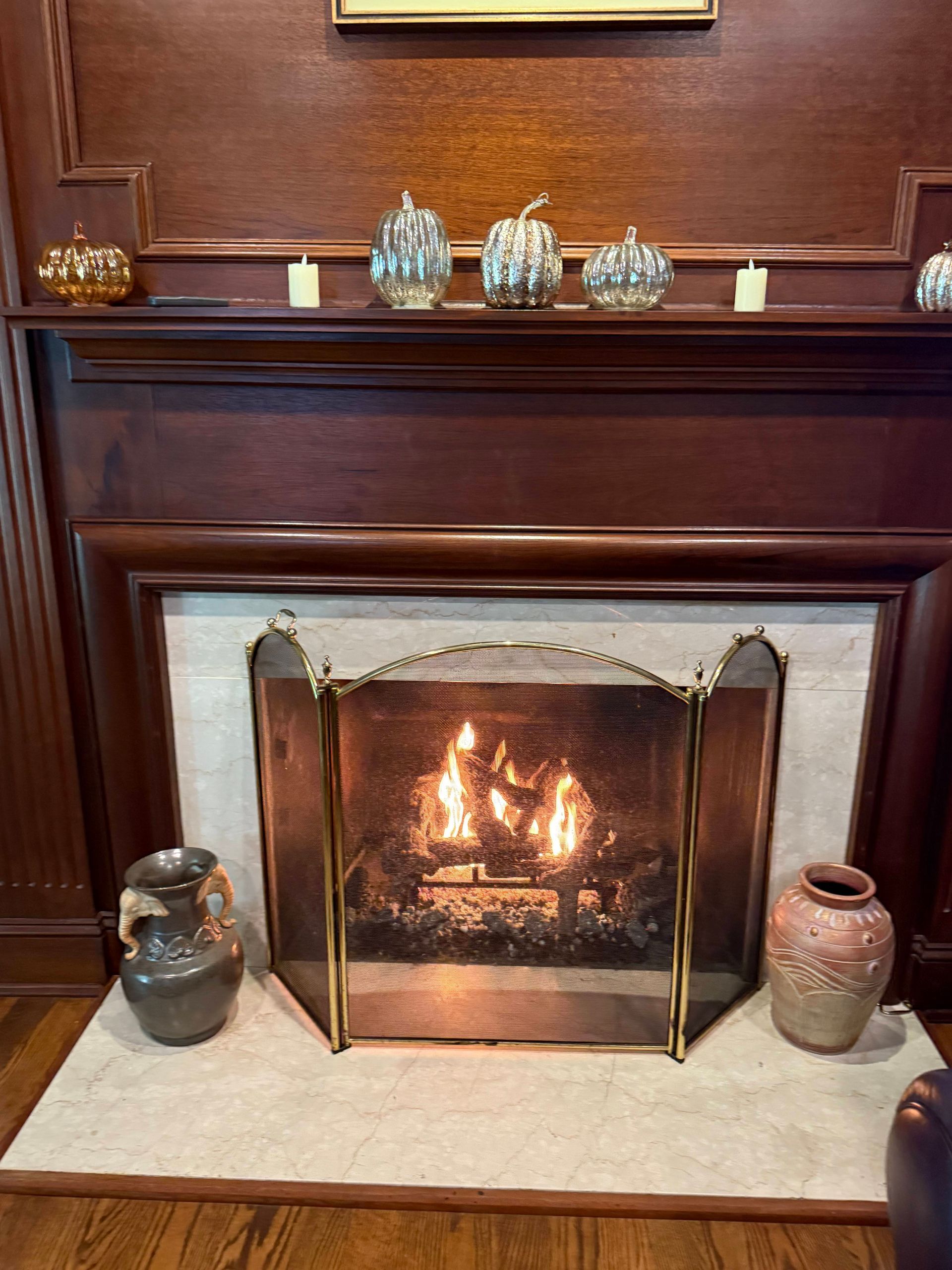 A fireplace with a lit fire, decorative glass pumpkins on the mantel, and two large vases on the stone hearth.