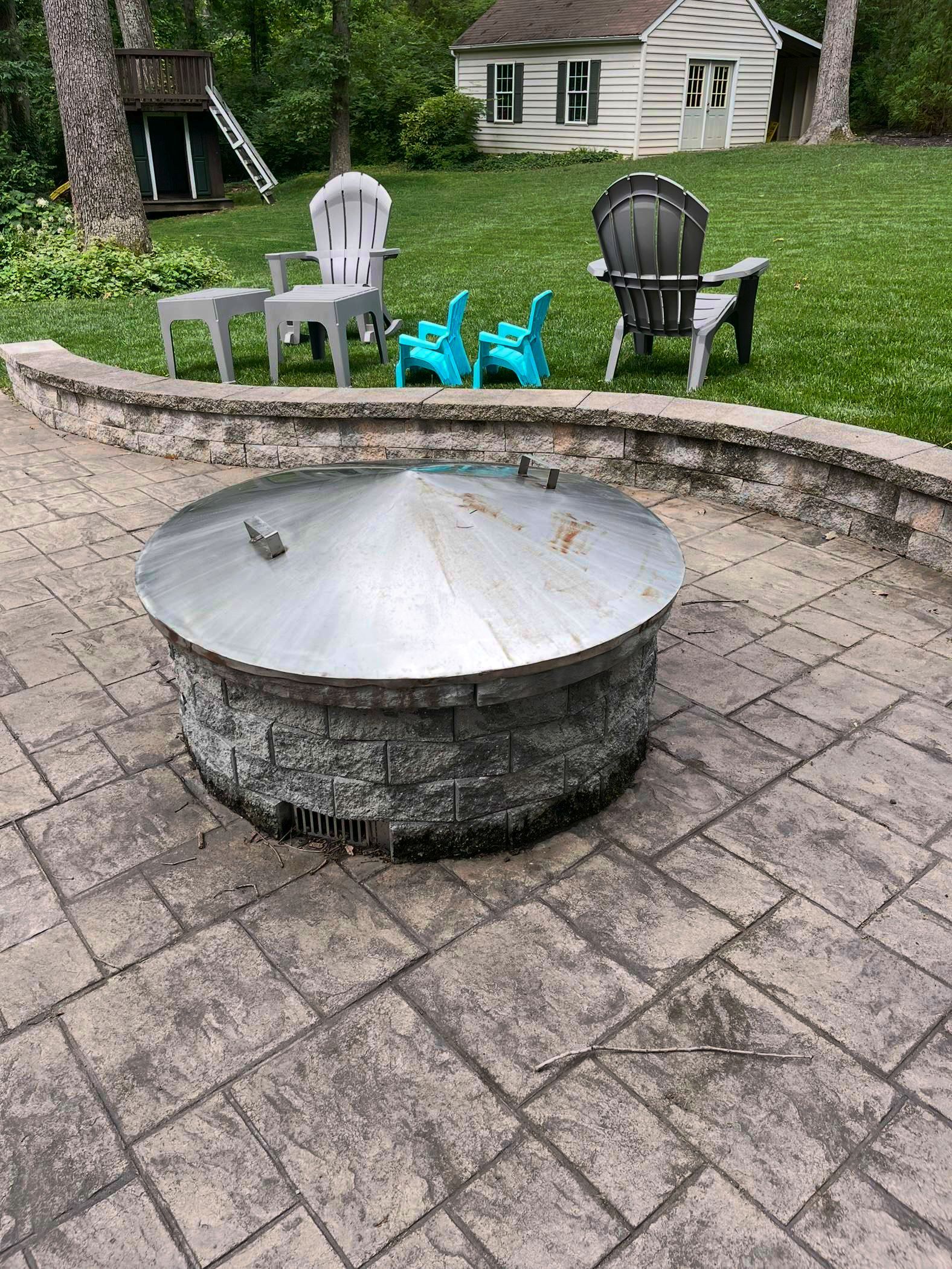 A stone fire pit with a metal lid on a paved patio, with outdoor chairs and a small white house in the background.