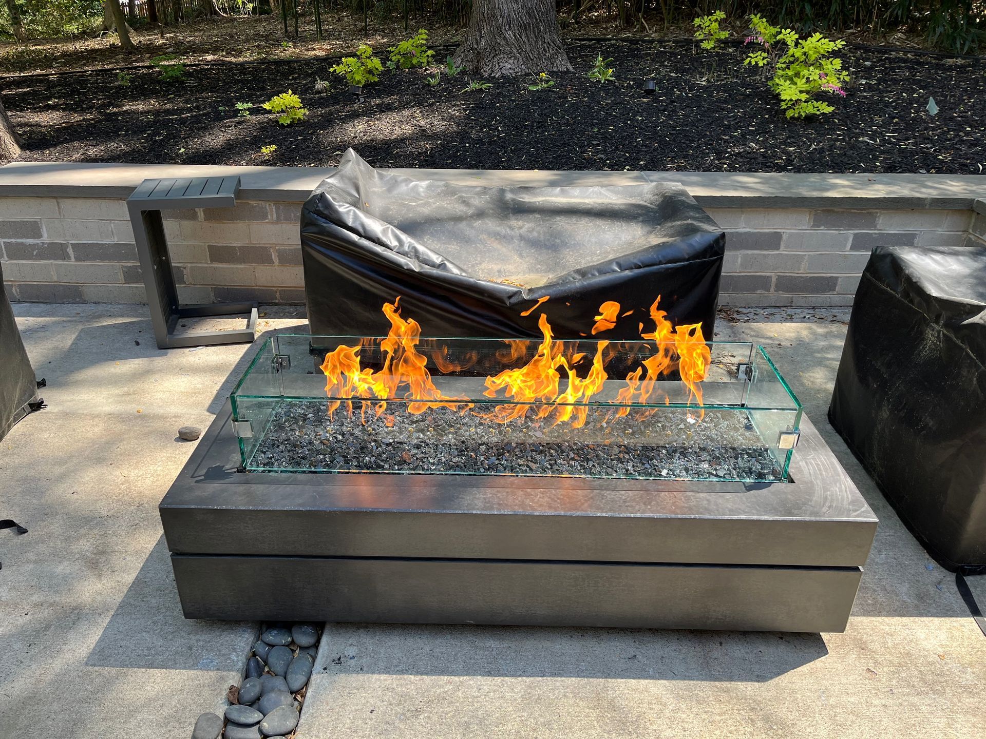 A modern, rectangular outdoor gas fire pit with bright flames, glass guards, and grey stone, set on a patio.