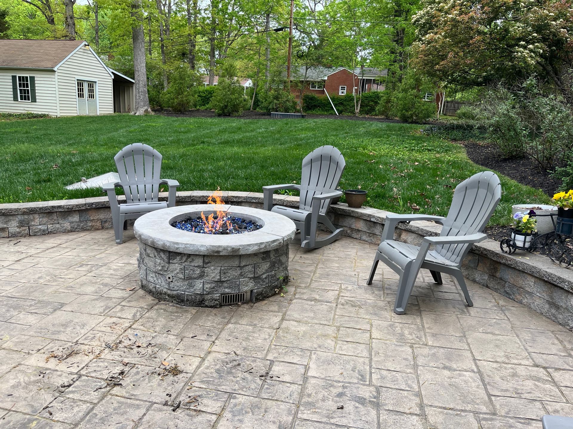 Three gray Adirondack chairs arranged around a circular stone fire pit on a patterned stone patio in a grassy backyard.