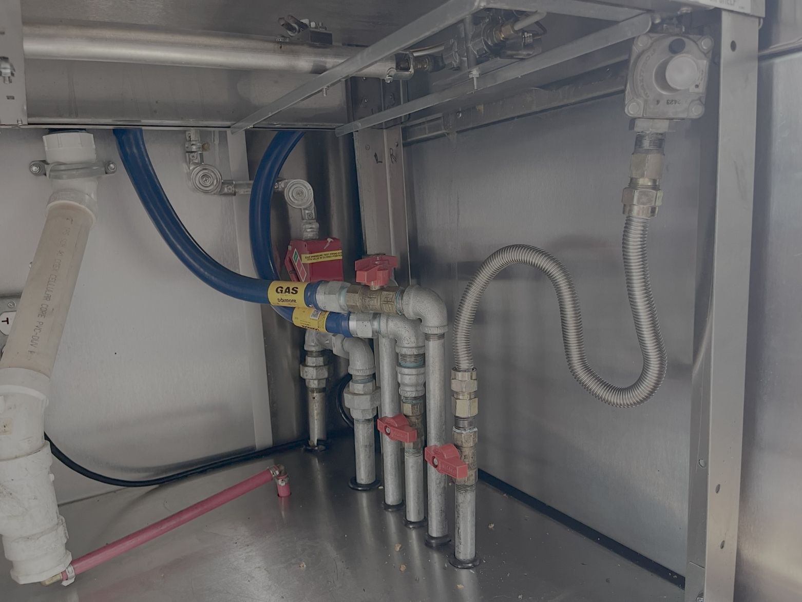 Under-sink plumbing with pipes, valves, and gas line in a commercial kitchen setting.