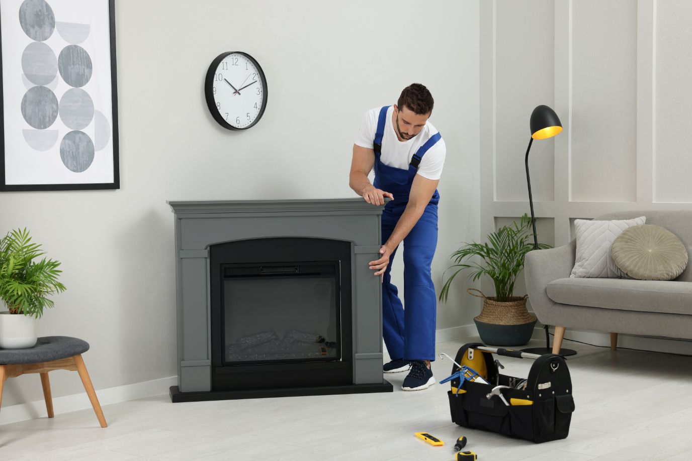 Man in blue overalls repairs a gray gas fireplace in a living room; a toolbox is on the floor.