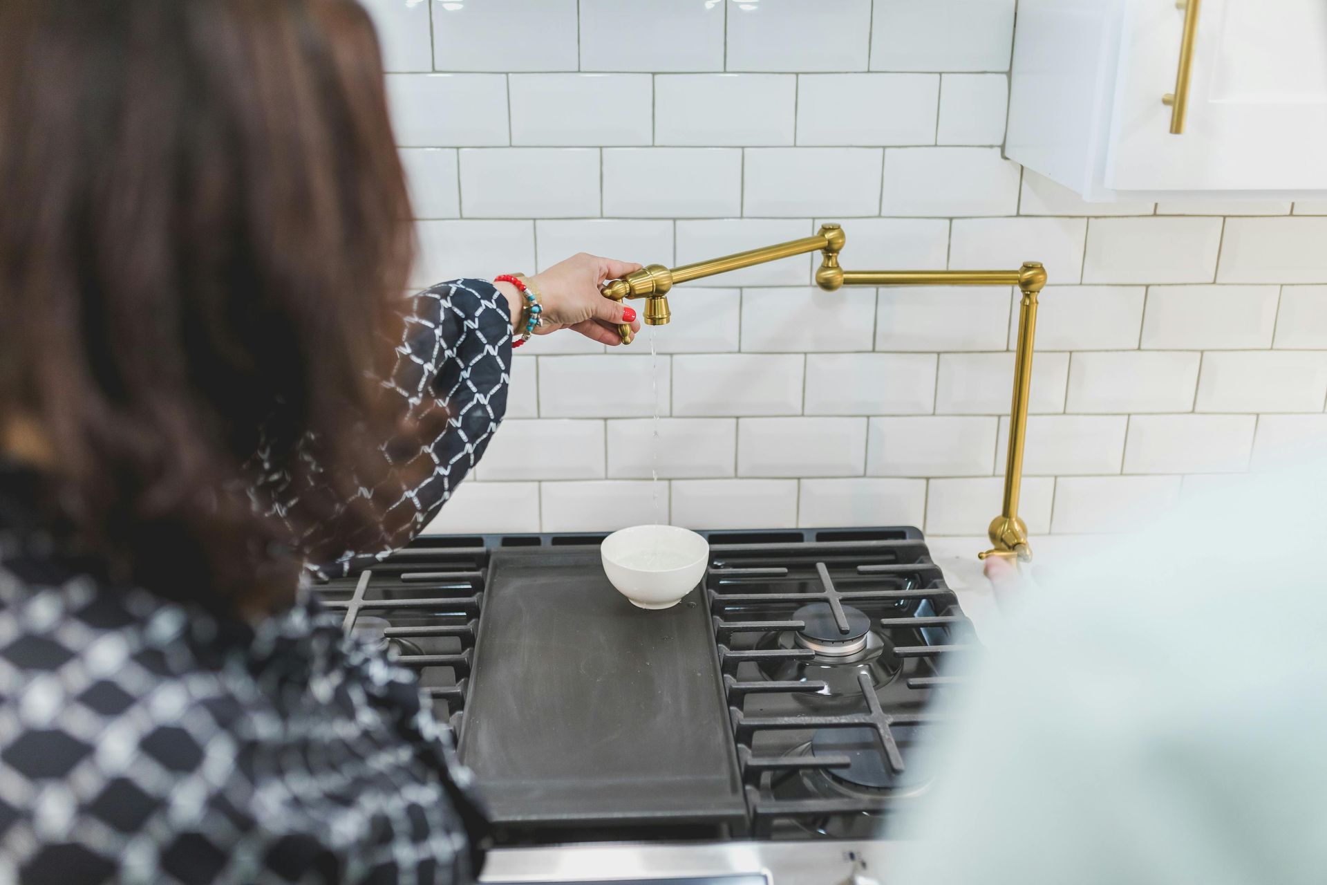 A person adjusts a brass wall-mounted pot filler faucet over a kitchen stove with a small white bowl on the grates.