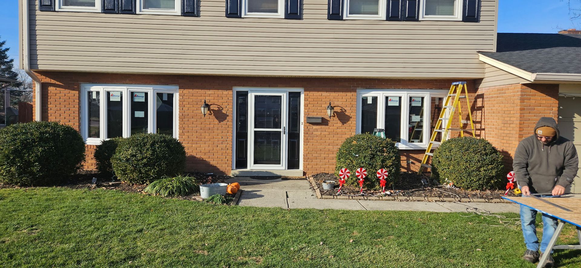 A man is standing in front of a brick house cutting a piece of wood.