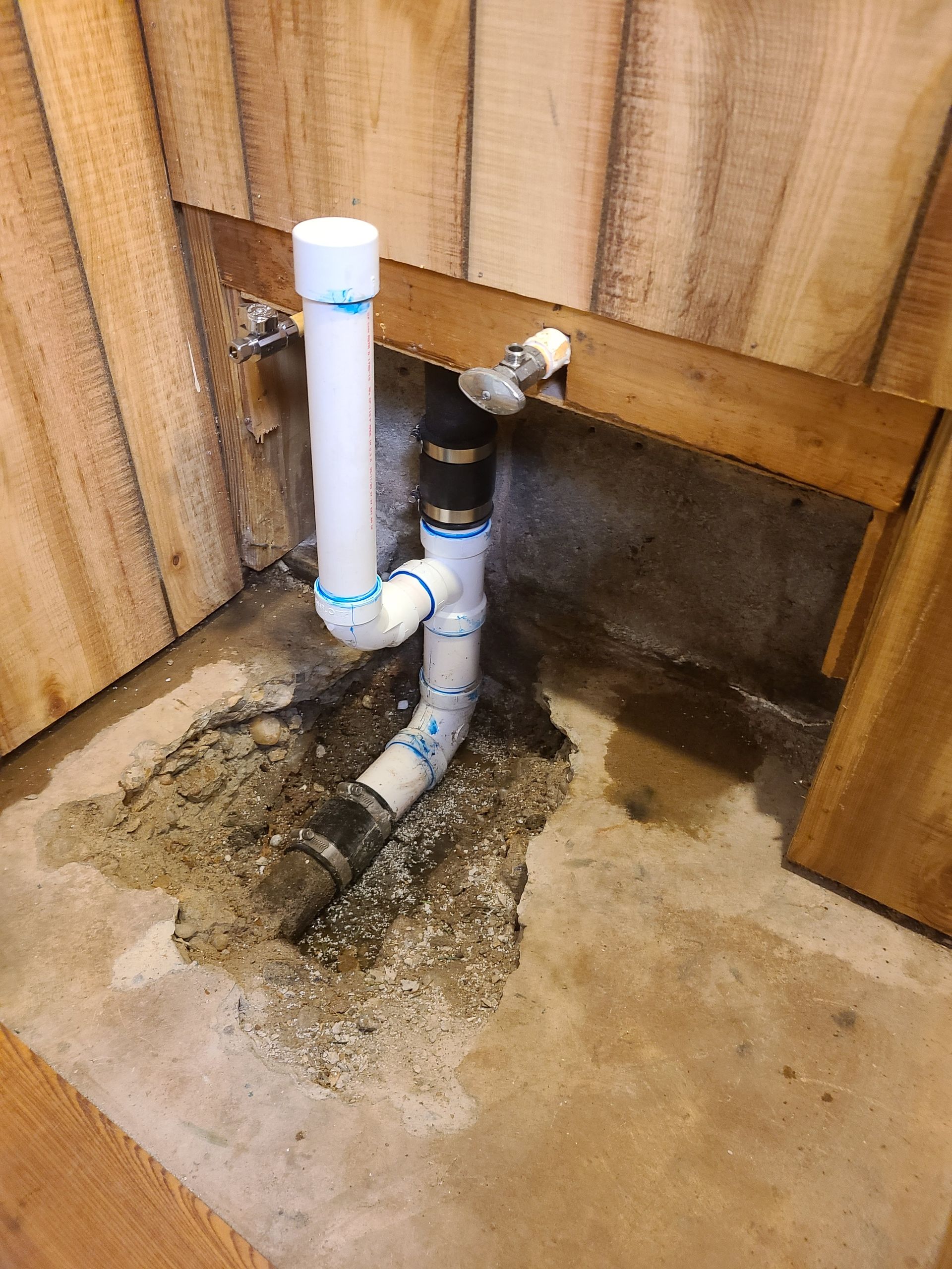 Plumbing pipes emerge from concrete floor, behind wooden wall. White and black pipes connected.