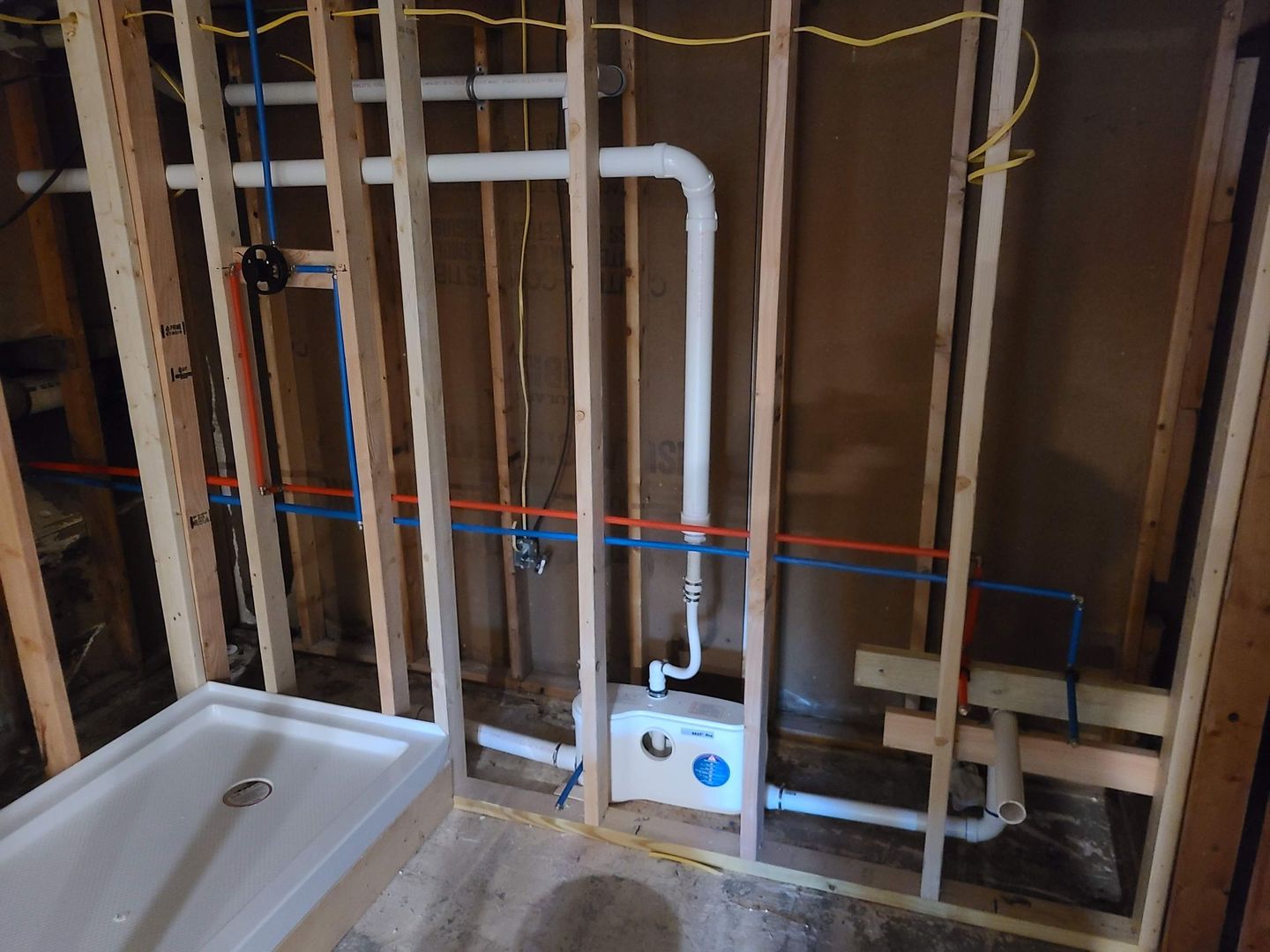 Bathroom under construction. Wood framing, white shower base, pipes, and electrical wiring visible.