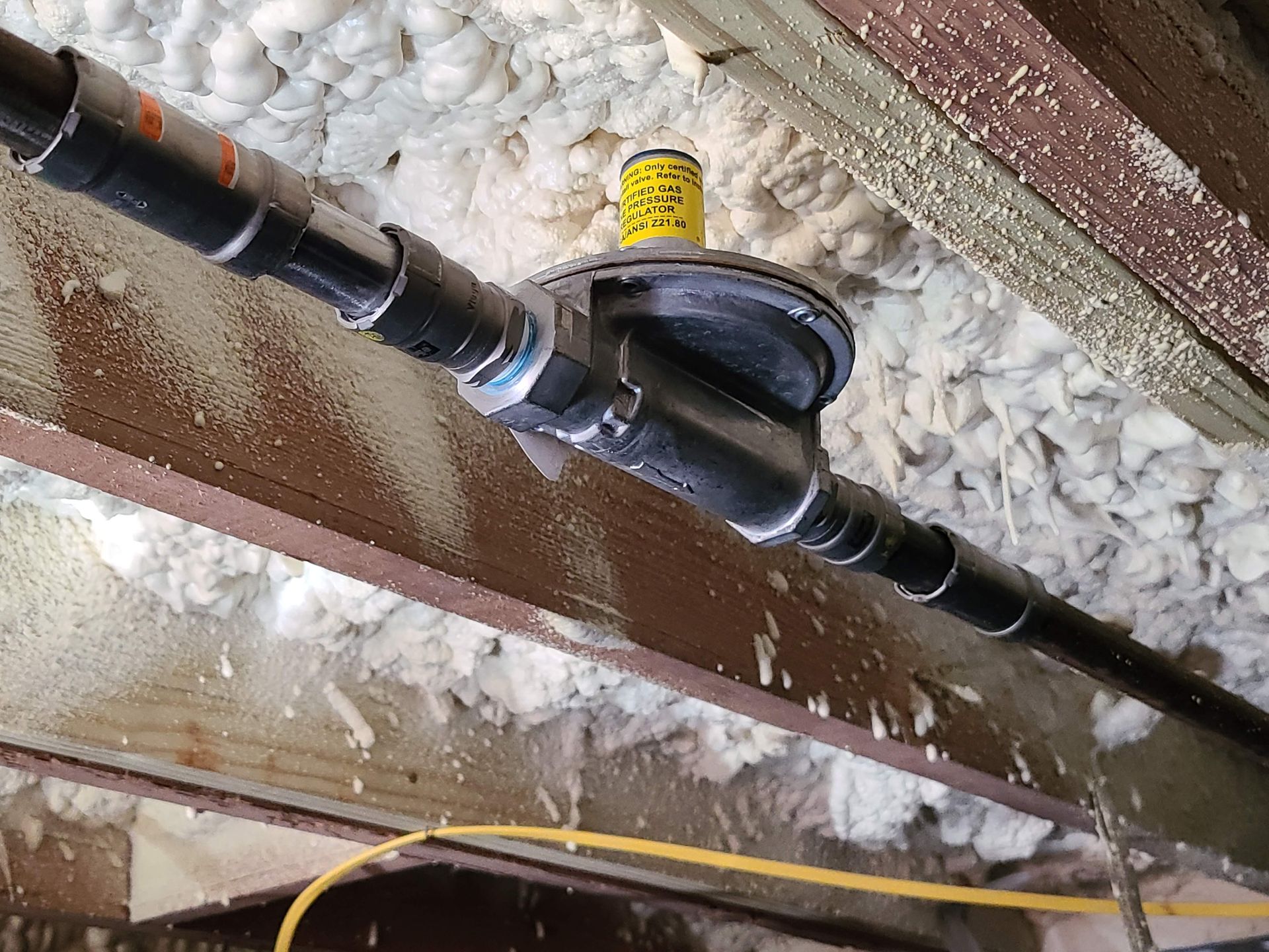 Black plumbing pipes and a black drain fitting are installed in a wooden space, near yellow insulation.