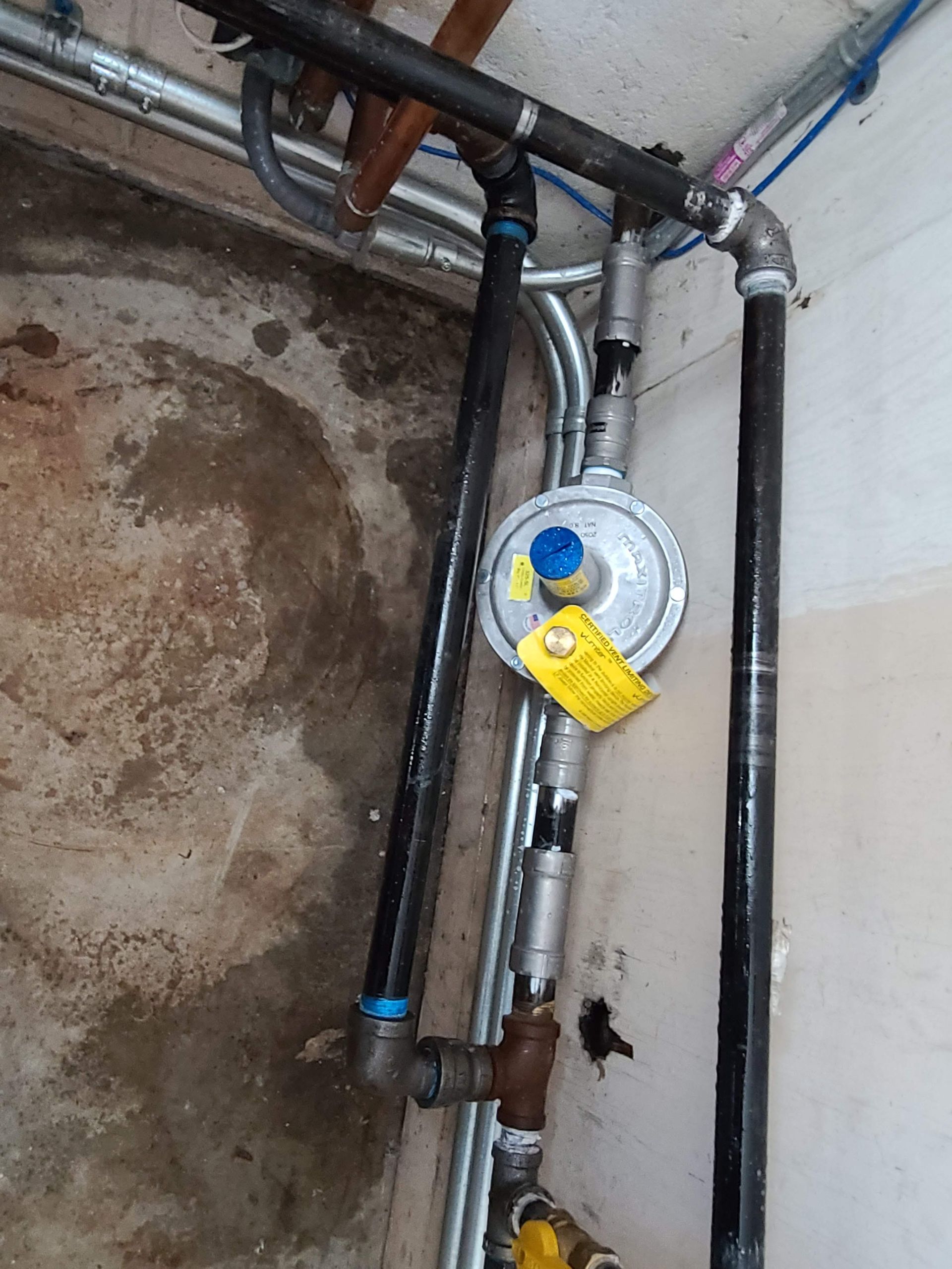 Gas regulator and piping in a utility area. Black and silver pipes, yellow tag.