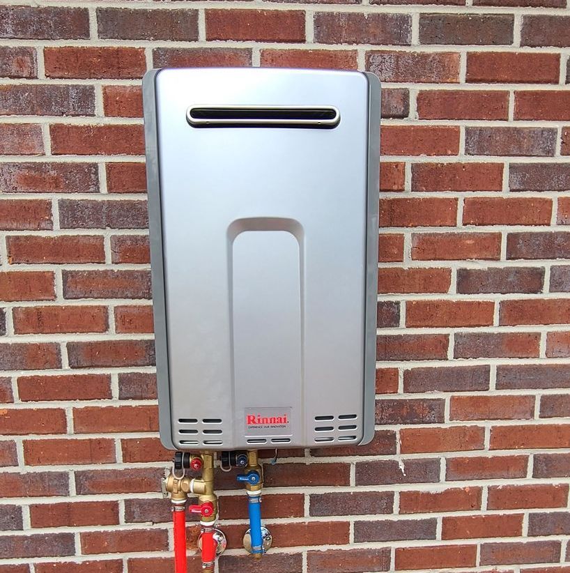 A silver, rectangular tankless water heater mounted on a red brick exterior wall.