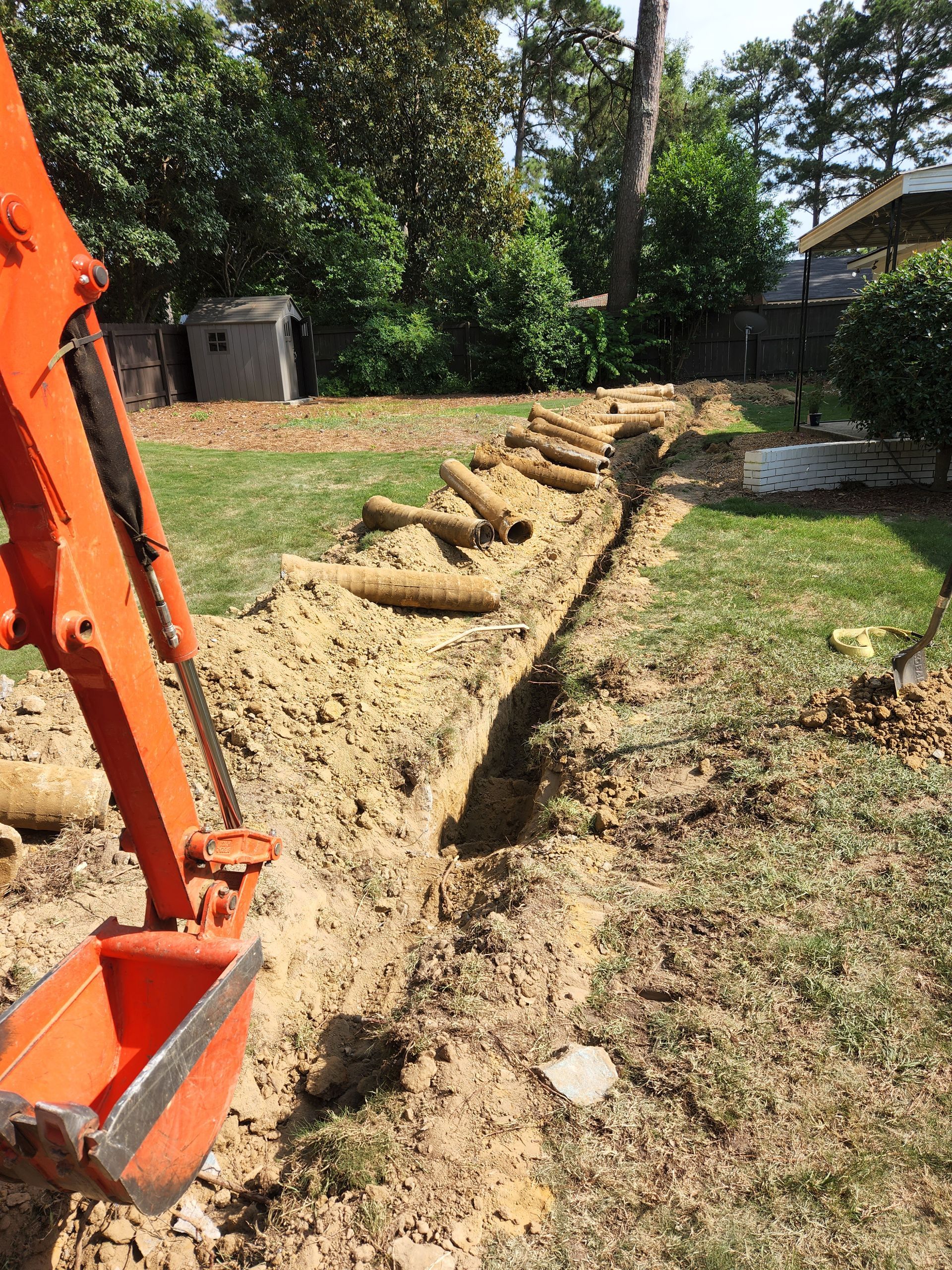 An orange excavator digging a trench in a grassy yard, preparing to lay pipes.