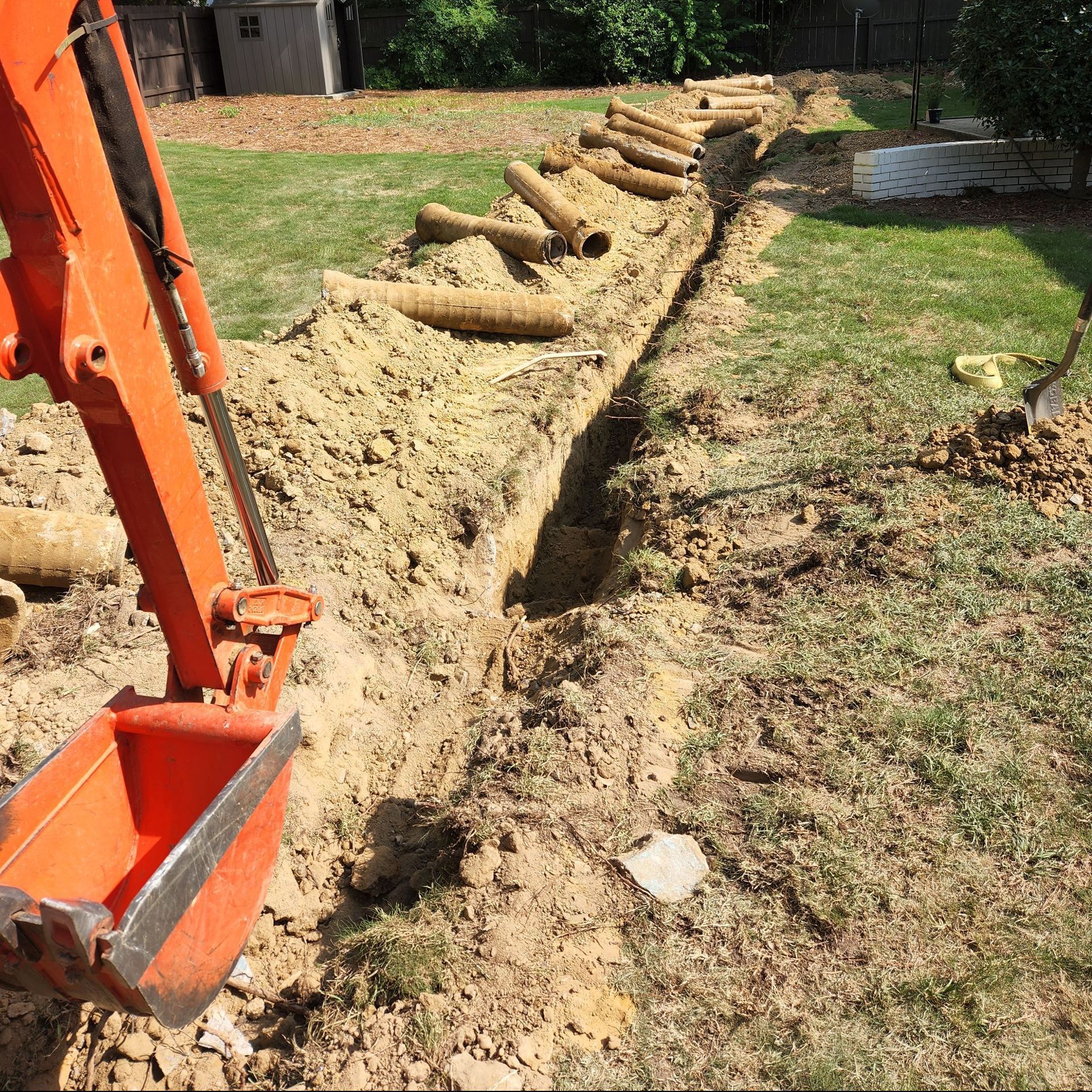 Orange excavator digging a trench in a yard; pipes lie alongside the trench.
