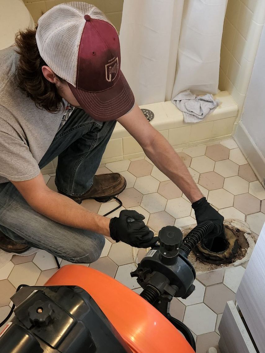 A person with a trucker hat unclogs a toilet drain in a bathroom with hexagon tile.