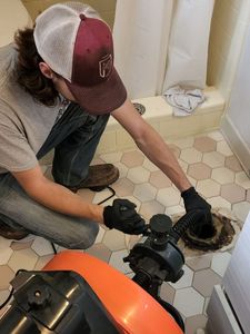 A person with a trucker hat unclogs a toilet drain in a bathroom with hexagon tile.