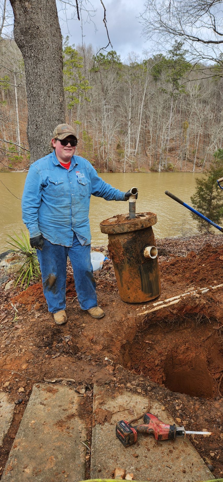 A man in denim stands near a well, holding the top. He stands near a body of water.